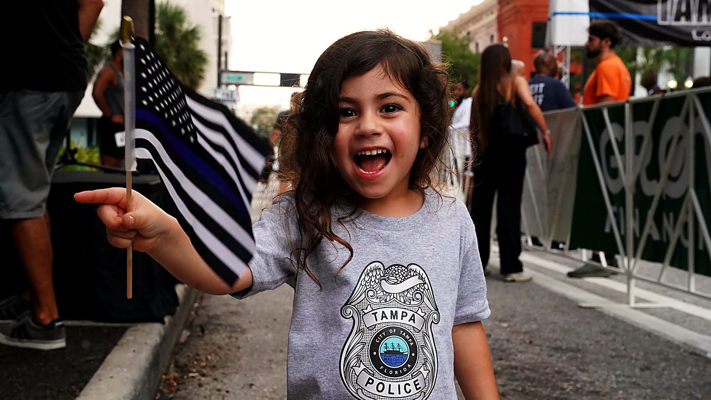 A young girl in a Tampa Police badge shirt waving a thin blue line flag at the run
