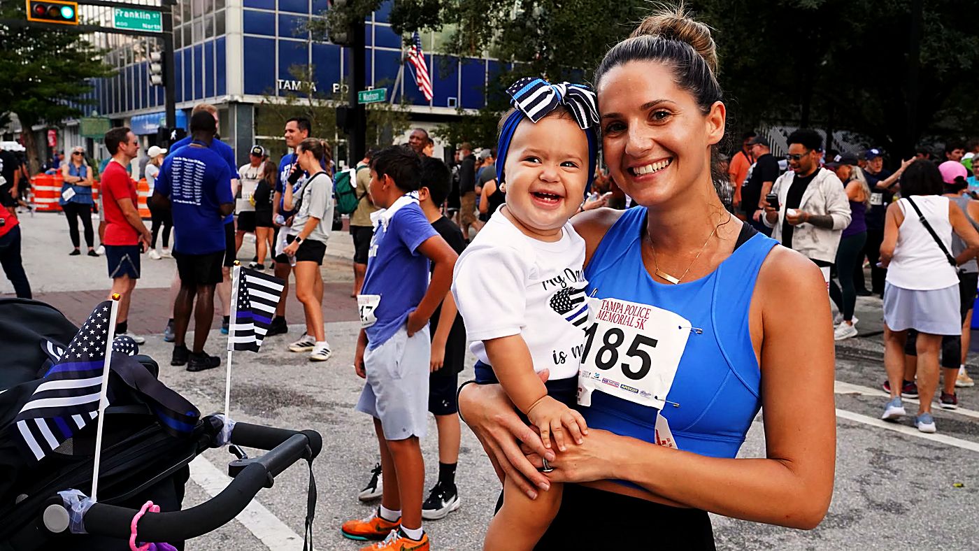 A runner holding her smiling baby, who wears a thin blue line headband, near the start line